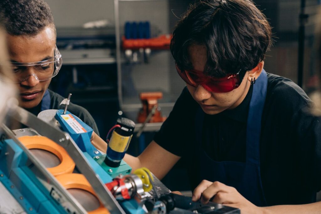 Two young engineers concentrating on precision work in a technical workshop, wearing safety goggles.